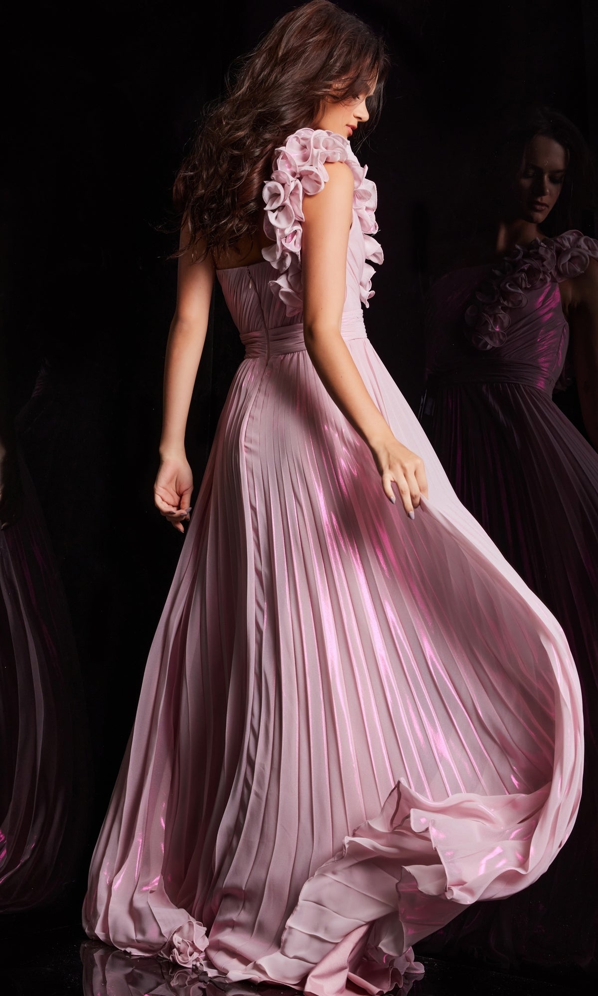 A woman is shown from the back against a dark background wearing the Jovani Long Pleated Chiffon Prom Dress 24609, featuring a flowing silhouette and floral details on the shoulders.